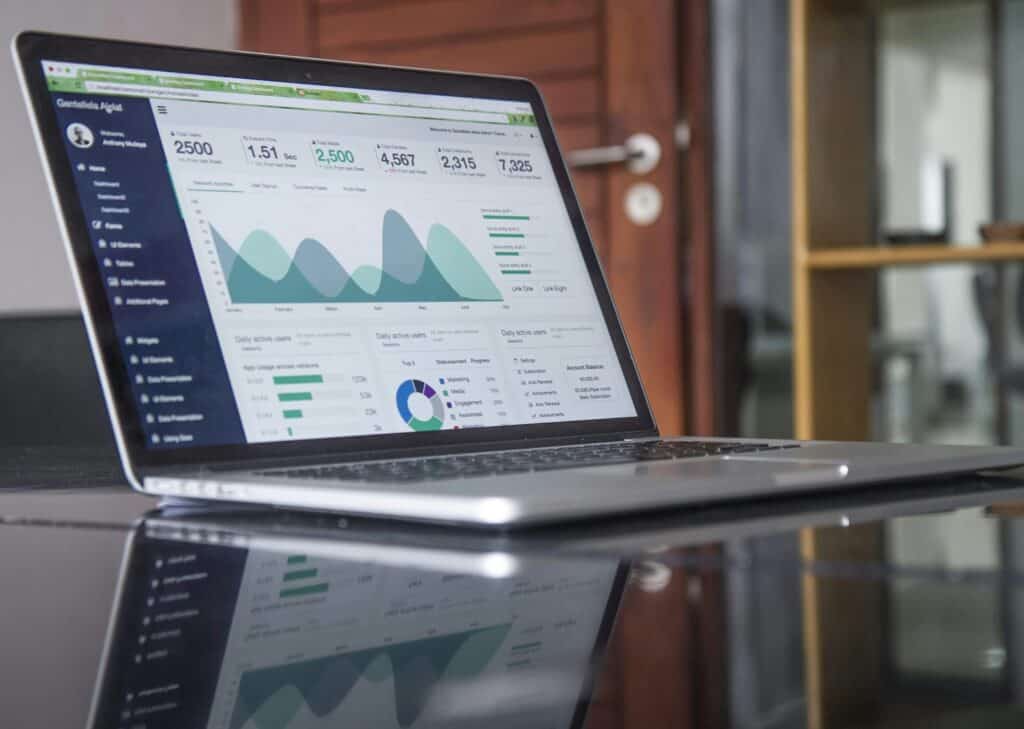 A laptop displaying a dashboard with various graphs and metrics, including line charts, bar charts, and numerical data. The screen shows analytics related to performance, such as visitor statistics and engagement levels, on a reflective surface in a professional setting.